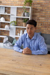 Asian man sitting at large wooden table in casual office, wearing light blue shirt, hands clasped