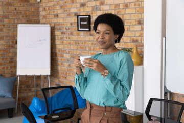 African American woman standing with white mug, wearing teal blouse in office near flip chart