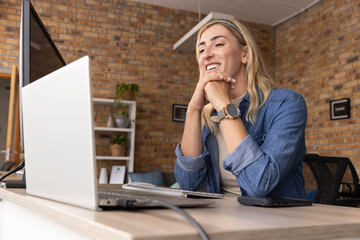 Mid adult woman in denim shirt over beige top, leaning at office desk with laptop, monitor