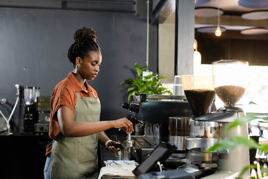 Adult African woman wearing green apron tamping portafilter at coffee counter with grinders
