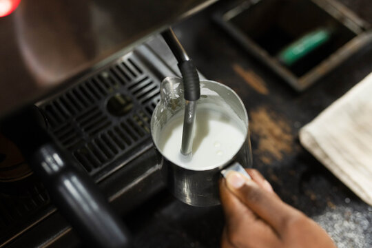 Stainless steel milk pitcher is being frothed by steam wand on worn counter near portafilter