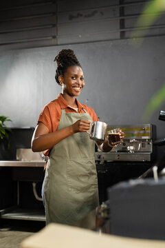 Adult woman smiling, preparing coffee at cafe counter, wearing olive apron and holding milk pitcher