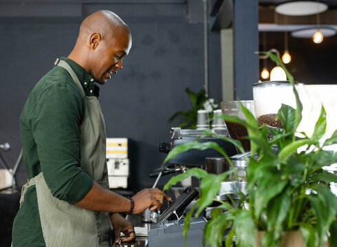 Adult African male in green shirt and apron operating register at cafe counter by espresso machine
