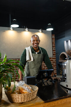 African American adult man leaning at wood counter in cafe, olive apron, towel, using POS, pastries