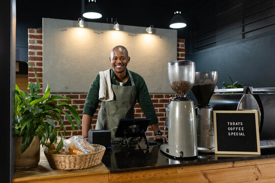Male barista wearing apron and towel standing behind wood counter in cafe using espresso machine