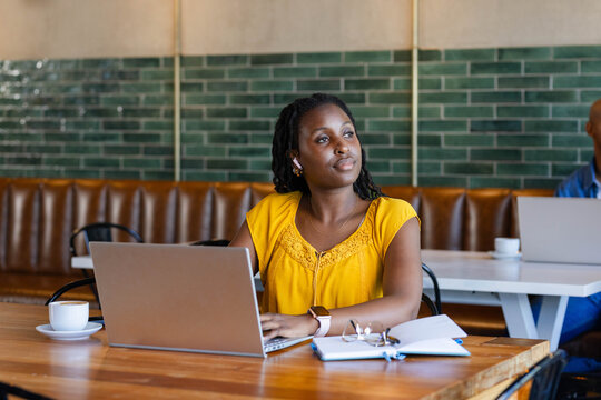 Adult African woman working on silver laptop at cafe wearing yellow blouse earbuds notebook resting