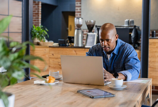 African American man working on laptop at cafe communal table with two white cups, wearing watch