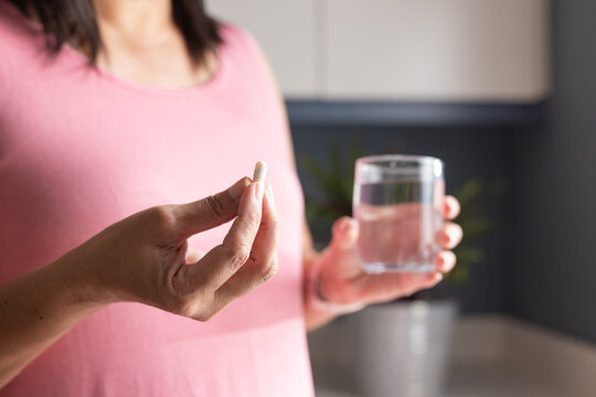 White capsule pill resting between fingers above clear glass of water on counter, soft blur