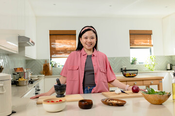 Asian woman wearing pink overshirt and headband preparing salad at modern kitchen island with knife