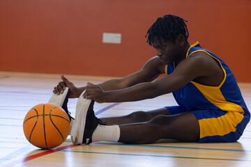 African American male athlete stretching on hardwood court, holding basketball and wearing uniform