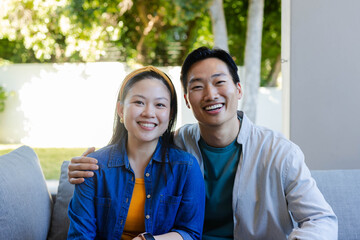 Asian couple sitting on light-gray sofa in covered patio, smiling at camera with yellow headband