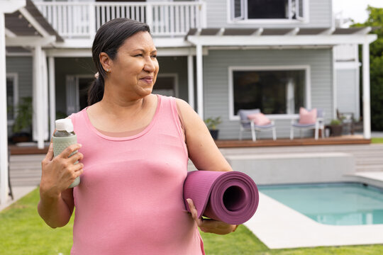 Woman wearing pink top standing by pool on lawn holding purple yoga mat and water bottle