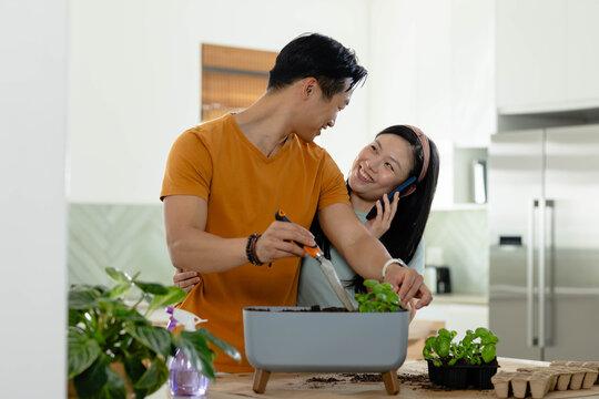 Asian couple potting herbs at kitchen counter with grey planter using orange trowel and smartphone