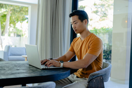 Asian man typing on silver laptop at table by glass doors in orange t-shirt with smartwatch