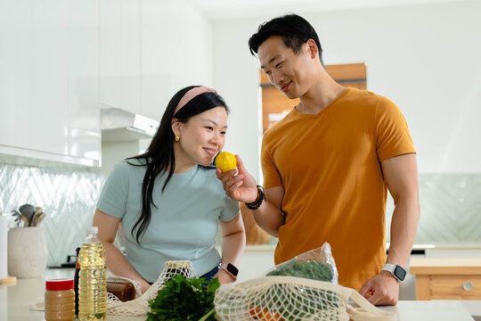 Asian couple unpacking groceries at kitchen island, holding lemon, inspecting mesh bags and herbs
