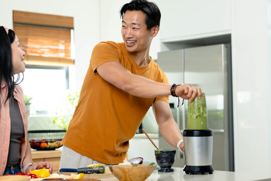 Asian couple preparing green smoothie in bright kitchen, man wearing mustard T-shirt using blender