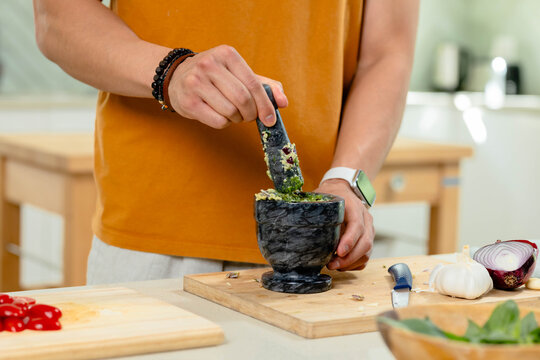 Dark marble mortar and pestle are crushing green herbs on wooden cutting board at kitchen island