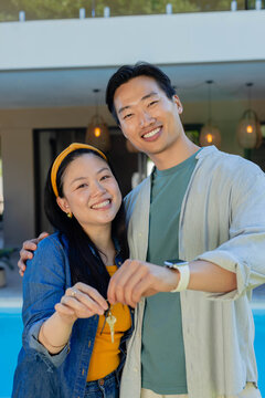 Asian couple smiling, holding house keys, posing by pool with denim jacket and smartwatch visible
