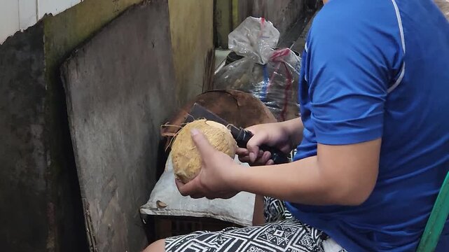 Worker opening hard coconut shell with knife for fresh preparation. Hands cracking mature coconut and separating meat in street kitchen. Manual coconut processing with blade in traditional market work