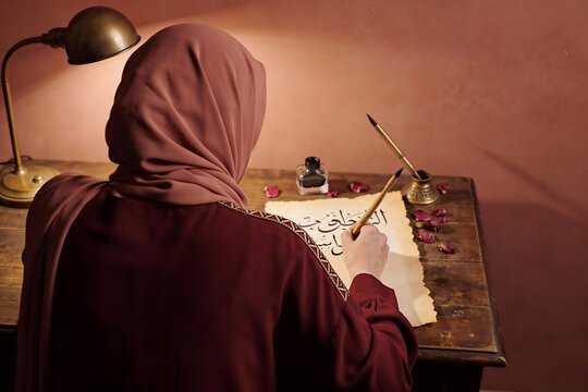 Rear view of woman in modest dress writing with traditional qalam pen on aged paper. Warm lamp light. Ideal for cultural arts, heritage content, creative lifestyle marketing materials.