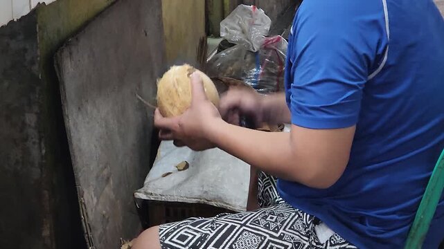 Man splitting coconut shell with knife during traditional food prep. Hands opening mature coconut and removing flesh in small workspace. Street vendor cracking hard coconut for fresh ingredient proces