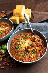 A rustic overhead view of homemade beef chili served in bowls and topped with shredded cheddar cheese, fresh herbs, and sliced jalape&ntilde;os. Surrounded by red beans, chili flakes, and cornbread on a wood