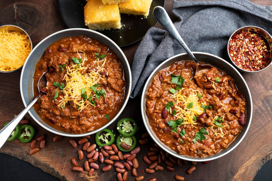 A rustic overhead view of homemade beef chili served in bowls and topped with shredded cheddar cheese, fresh herbs, and sliced jalape&ntilde;os. Surrounded by red beans, chili flakes, and cornbread on a wood