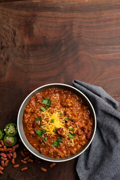 A rustic overhead view of homemade beef chili served in bowls and topped with shredded cheddar cheese, fresh herbs, and sliced jalape&ntilde;os. Surrounded by red beans, chili flakes, and cornbread on a wood