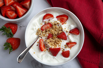 A bowl of creamy Greek yogurt topped with crunchy granola and fresh sliced strawberries, photographed in natural light on a marble surface. Styled as a healthy breakfast or snack with clean, minimal c