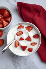 A bowl of creamy Greek yogurt topped with crunchy granola and fresh sliced strawberries, photographed in natural light on a marble surface. Styled as a healthy breakfast or snack with clean, minimal c