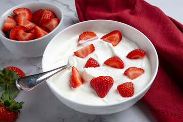 A bowl of creamy Greek yogurt topped with crunchy granola and fresh sliced strawberries, photographed in natural light on a marble surface. Styled as a healthy breakfast or snack with clean, minimal c