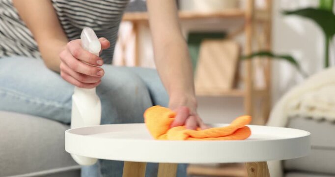 Woman spraying cleaning product and wiping table with microfiber cloth at home, closeup