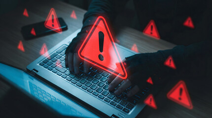 Anonymous hacker typing on a laptop keyboard surrounded by glowing red warning signs at night