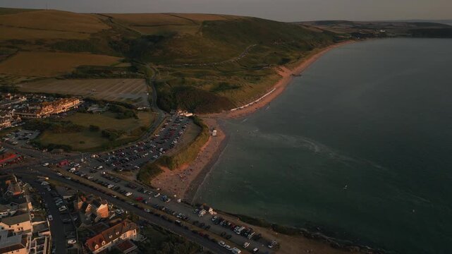 Coastal town and beach on a sunny day, with cars parked along the road and beachgoers enjoying the water and sand.