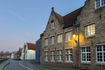 Bruges, Belgium – December 26, 2025. Historic Flemish brick houses with stepped gables and warm street lanterns at dusk, glowing windows and cozy evening atmosphere in the old town.