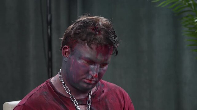 A man in theatrical zombie makeup, with simulated blood and bruises, and a heavy chain around his neck, sits with his head down, trying to escape against the backdrop of a dark room.