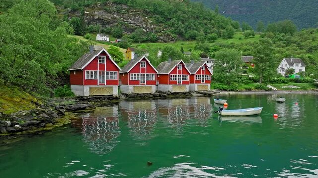 Red boathouses lining the fjord shoreline in Fl&aring;m, Norway beneath green mountains. Tranquil Norwegian village scene with calm water highlights rural life and nature.