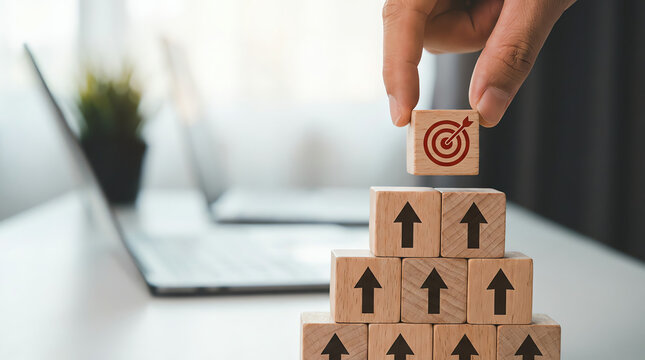 Achieving your goals by stacking wooden blocks to build a pyramid with a target on the top block and a person's hand placing it on top