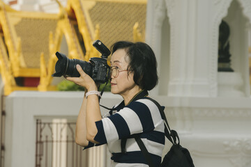 Woman with glasses and striped shirt is taking photo with DSLR camera in outdoor setting, possibly near temple. scene conveys focus and interest in photography