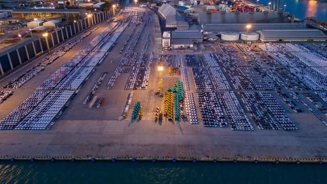 Aerial night view of a massive automotive port terminal filled with thousands of new cars ready for export. Industrial logistics landscape with rows of parked vehicles illuminated by floodlights 
