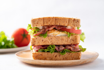 Whole wheat bread sandwich with bacon, lettuce, tomato and fried egg on wooden plate with white background