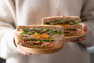 Whole wheat bread sandwich with bacon, lettuce, tomato and fried egg on wooden plate holding by woman hand