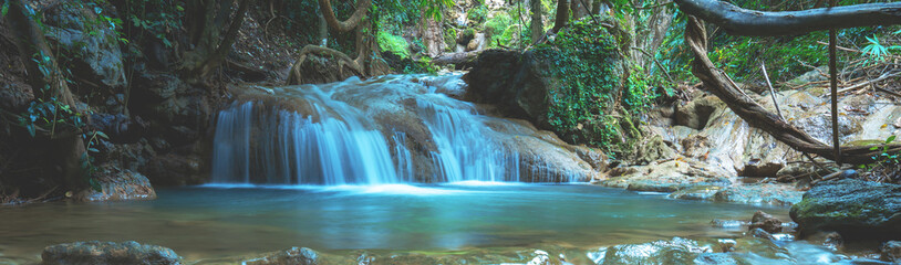 Stunning panoramic landscape of a serene waterfall cascading into a clear blue pool in a lush tropical forest. Wide horizontal nature banner background with silk water flow and mossy rocks.