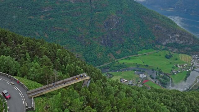 Drone view from Stegastein overlooking Aurlandsfjord and valley below in Norway. Green hills, village, and shoreline appear far beneath the mountain road and viewpoint.