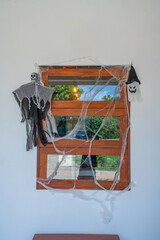 Halloween home window decoration featuring artificial spider webs, a hanging skeleton ghost, and spooky ornaments on a wooden frame. Creative exterior festive decor for October celebrations.