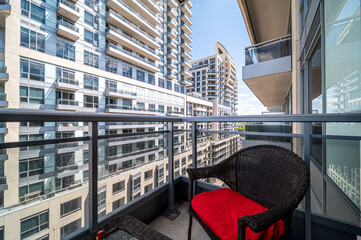 Obraz premium Balcony featuring a wicker chair with a red cushion, offering a view of contemporary high rise buildings and the expansive urban environment, embodying city living