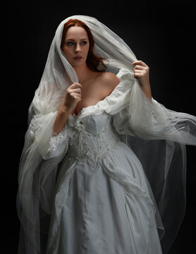 close up portrait female model wearing antique white historical ghost bride wedding dress gown, ethereal bridal veil head covering. Isolated standing figure, dark studio background overhead lighting