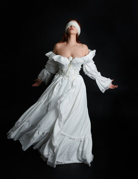 full length portrait beautiful female model wearing antique white historical ghost bride wedding dress gown, veiled mask blindfold covering face. isolated walking figure on dark studio background.
