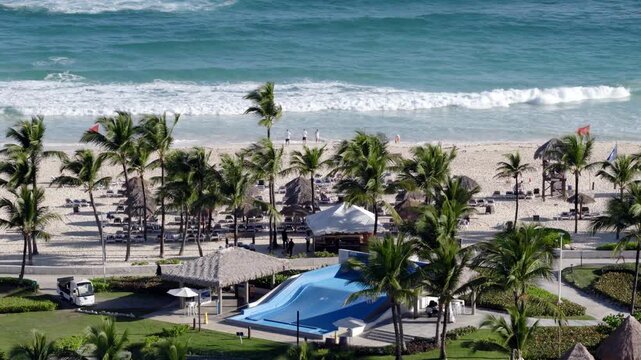 Aerial view of strong winds along Punta Cana coastline, with palm trees bending near Hard Rock Hotel beachfront and rolling waves on Caribbean shore. Sandy beach of famous hotel. Wide shot.