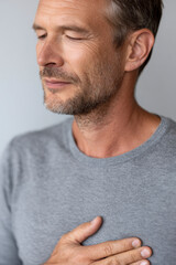 Obraz premium Portrait of a man in a grey shirt with his eyes closed and his hand over his heart in gratitude.